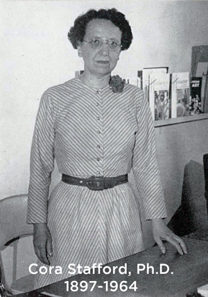 Cora Stafford standing at her desk, circa 1950s, short dark hair, glasses, striped dress