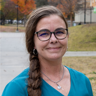 Kim Willis facing forward, smiling, long braided hair, wearing glasses, earrings and a turquoise-colored top